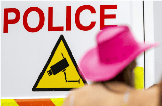 Back of a woman in a pink cowboy hat in front of a sign that says police in red letters with a yellow icon of a surveillance camera